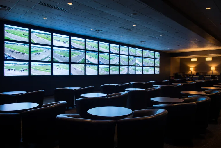 Illustration: a dark, empty lounge with rows of chairs and small tables facing a wall of many screens, each showing a distant racetrack.